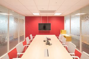 Une salle de conférence moderne avec une longue table en bois, huit chaises, une télévision murale et un mur d'accent rouge. Les murs de la salle sont des cloisons vitrées avec des motifs givrés et un éclairage lumineux.