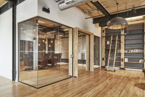 Espace de bureau moderne avec salle de réunion vitrée, sols en bois, plafond industriel et grande étagère.